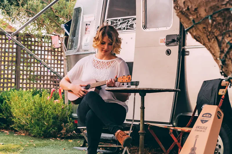 woman playing a Kala ukulele while sitting outside a travel trailer surrounded by nature