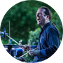 A circular cutout photo of a Erik Kaszynski drumming with long hair, wearing a dark blue denim shirt with a green foliage background.