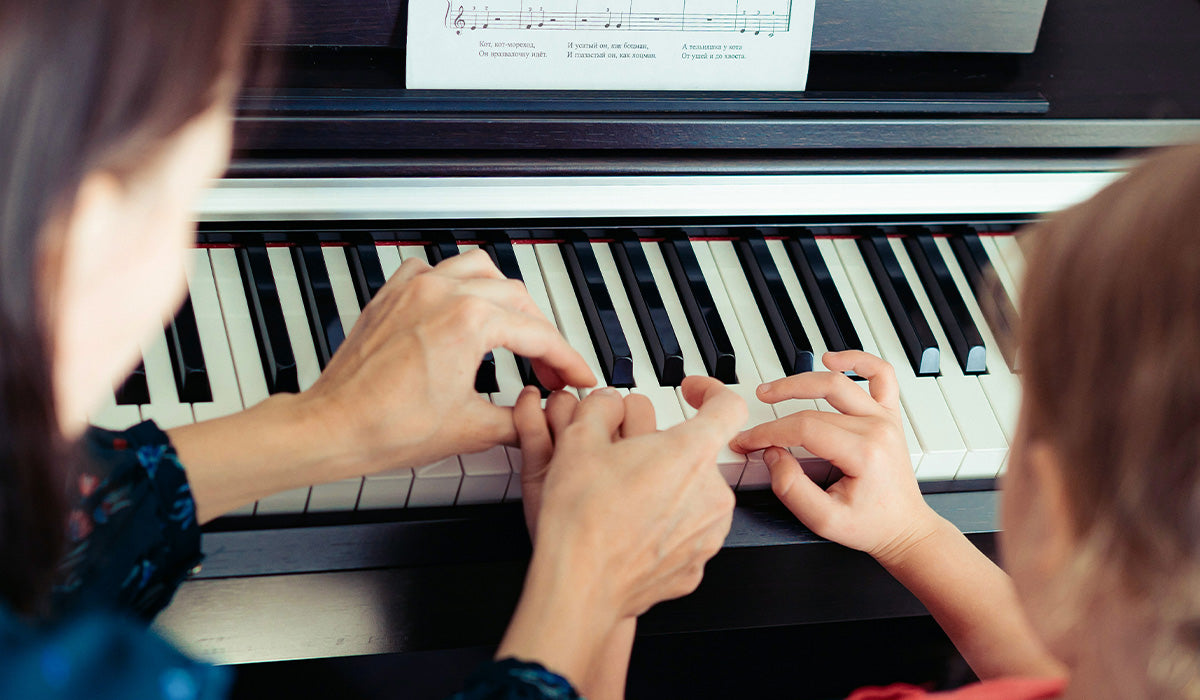 A close-up view of a person and a child playing a black digital piano, with hands pressing the keys and sheet music visible on the stand.