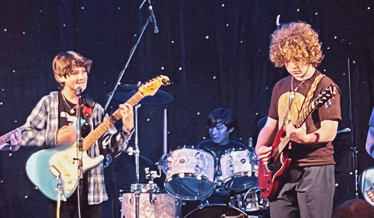 Two young musicians performing on stage, one playing a light blue electric guitar and the other playing a red electric guitar, with a drummer visible in the background against a dark starry curtain.