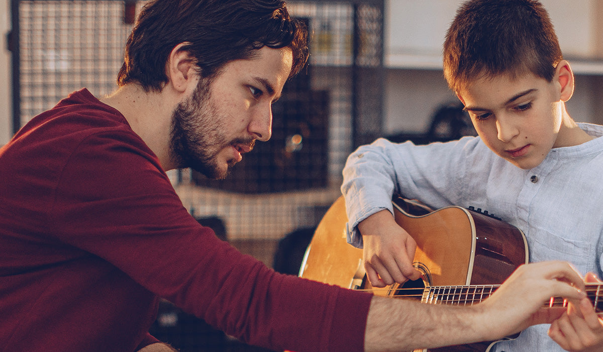 A man in a burgundy long-sleeve shirt teaches a young boy how to play an acoustic guitar in a warmly lit indoor setting.