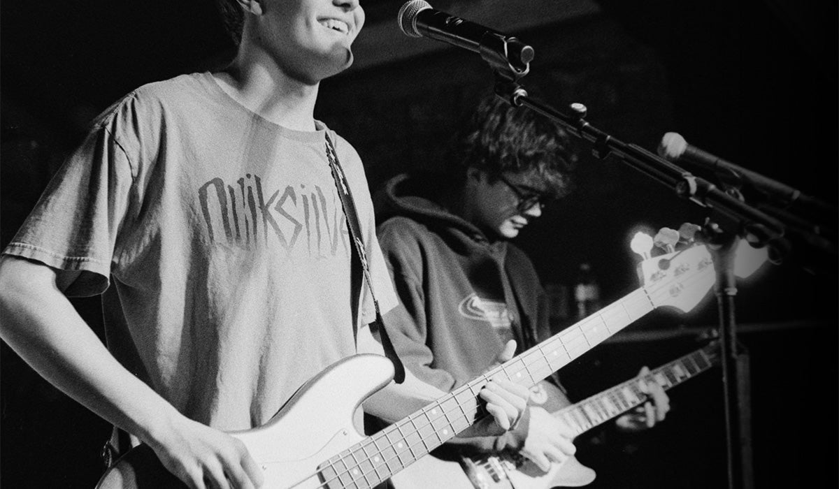 Black and white photo of a band performing on stage, featuring a smiling guitarist in the foreground wearing a Quiksilver t-shirt and playing a white electric guitar, with a bassist behind him near a microphone.