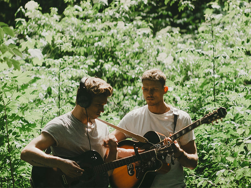 Two musicians playing acoustic guitars outdoors in wooded setting