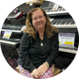 Photo of Marylee Logan Fairfield with glasses and shoulder-length wavy hair sitting in a music store surrounded by keyboards.