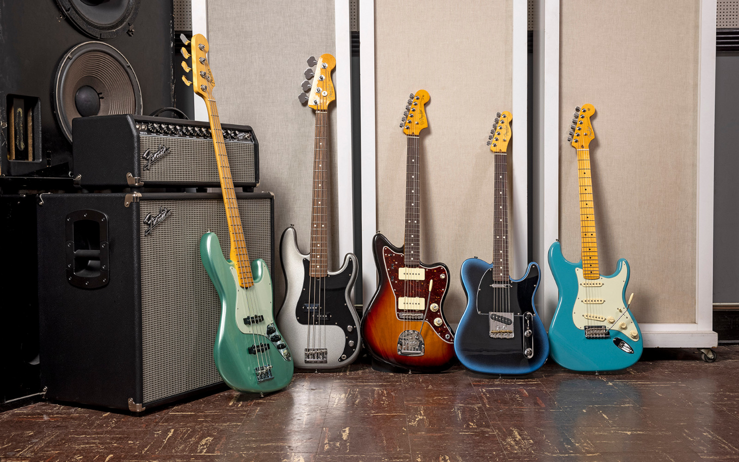 Five Fender American Pro II electric guitars and a bass guitar displayed upright in a studio setting, with large amplifiers on the left. The guitars include mint green, white, sunburst brown, dark blue, and turquoise finishes, with yellow and maple necks.