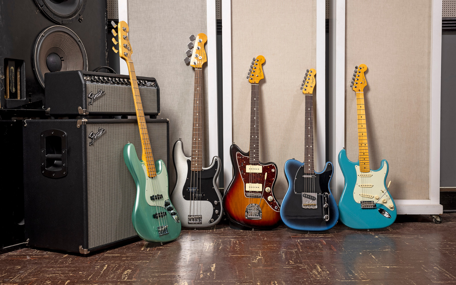 Five Fender American Pro II electric guitars and a bass guitar displayed upright in a studio setting, with large amplifiers on the left. The guitars include mint green, white, sunburst brown, dark blue, and turquoise finishes, with yellow and maple necks.