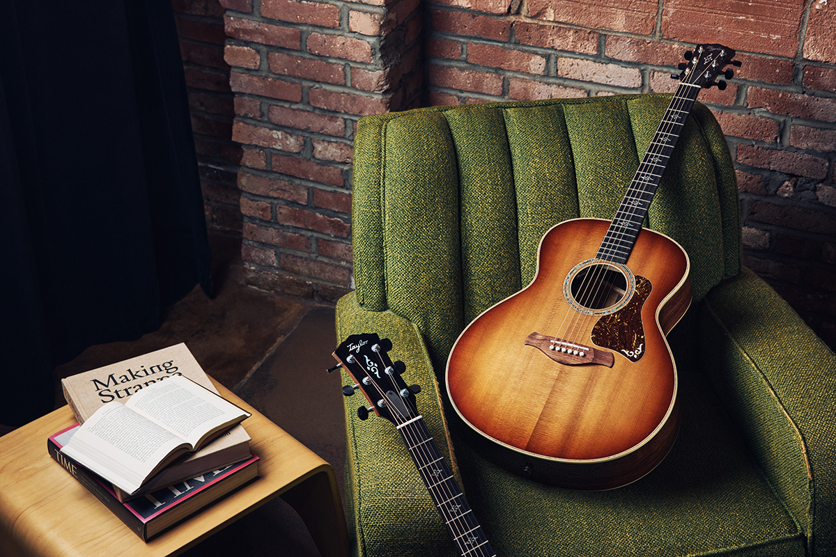 Acoustic guitar leaning against a green armchair with an open book on a small table in front of a brick wall.