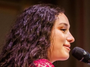 A close-up side profile of a woman with long, curly dark hair speaking into a black microphone. She is wearing a bright pink top
